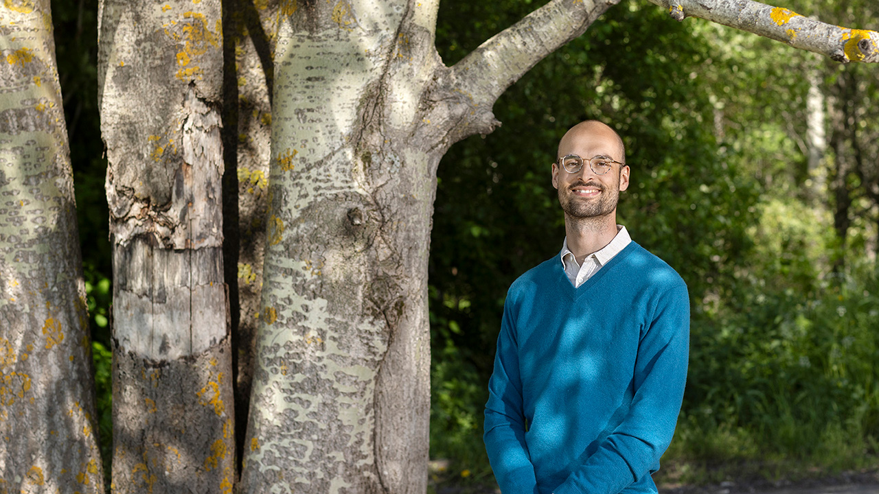 Portrait of a man with a blue sweater who is standing next to a tree. He has glasses and short hair and is smiling into the camera.