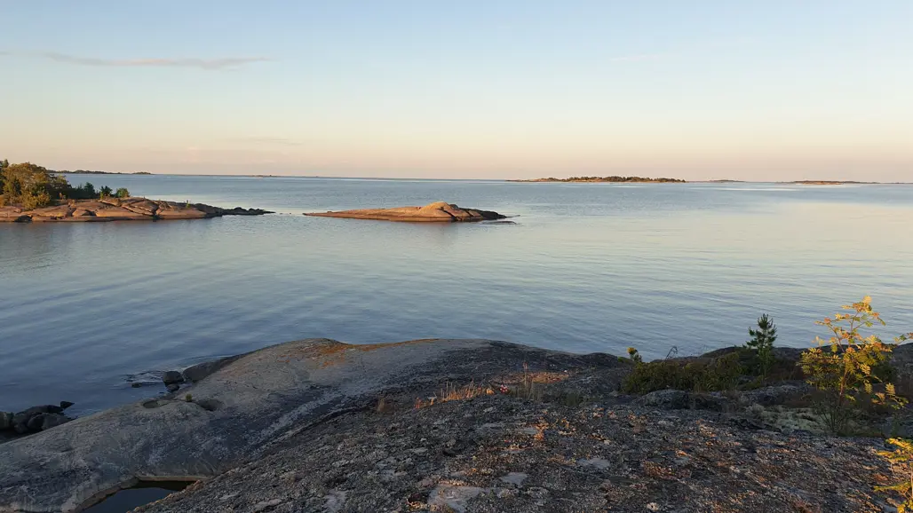 Kustlandskap i kvällsljus med släta klippor och stilla vatten, små öar i bakgrunden och låg vegetation i förgrunden