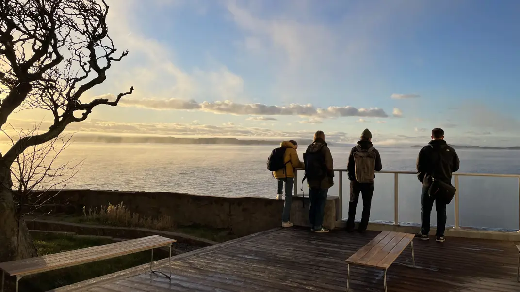 Four people are standing by a railing on a wooden veranda, looking out over an ocean or a large lake. The sun is setting or rising, creating a golden light on the water and sky. A crooked tree branch is visible on the left side of the picture, and two simple benches are on the porch. It is a chilly day, as the people are wearing warm jackets and backpacks.