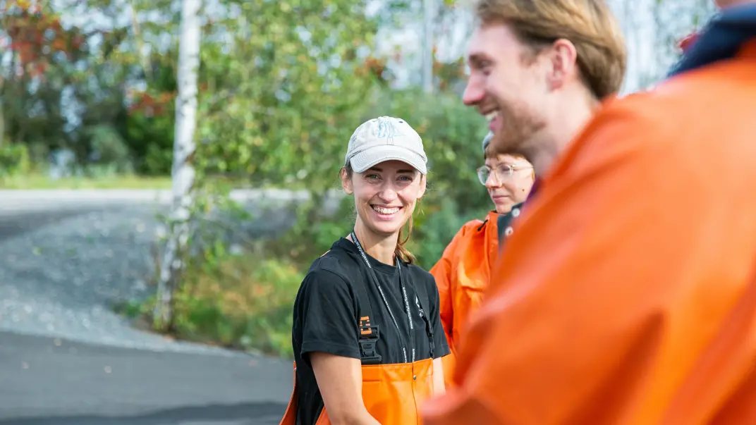 A smiling person in a cap and orange apron stands outside looking at the camera. In the background are more people in similar clothing, with trees and a dirt road in the background