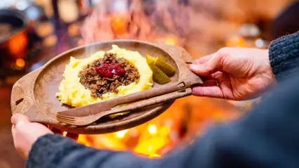 En person håller upp en tallrik med potatismos och renkött. Eldiga färger i bakgrunden.