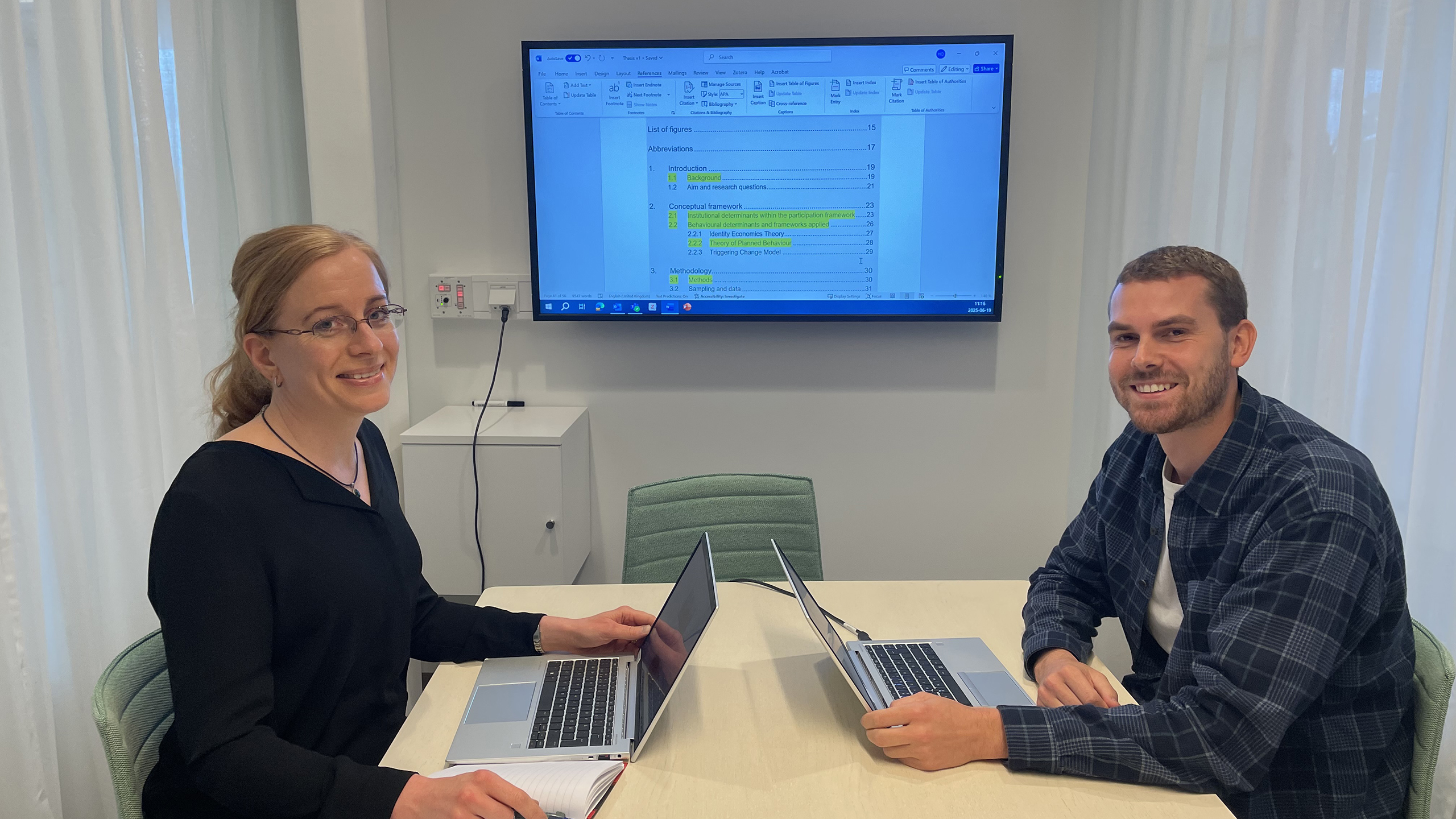 Two persons sitting by a table in an office looking into the camera. On the wall behind them a monitor showing a word processing program. 