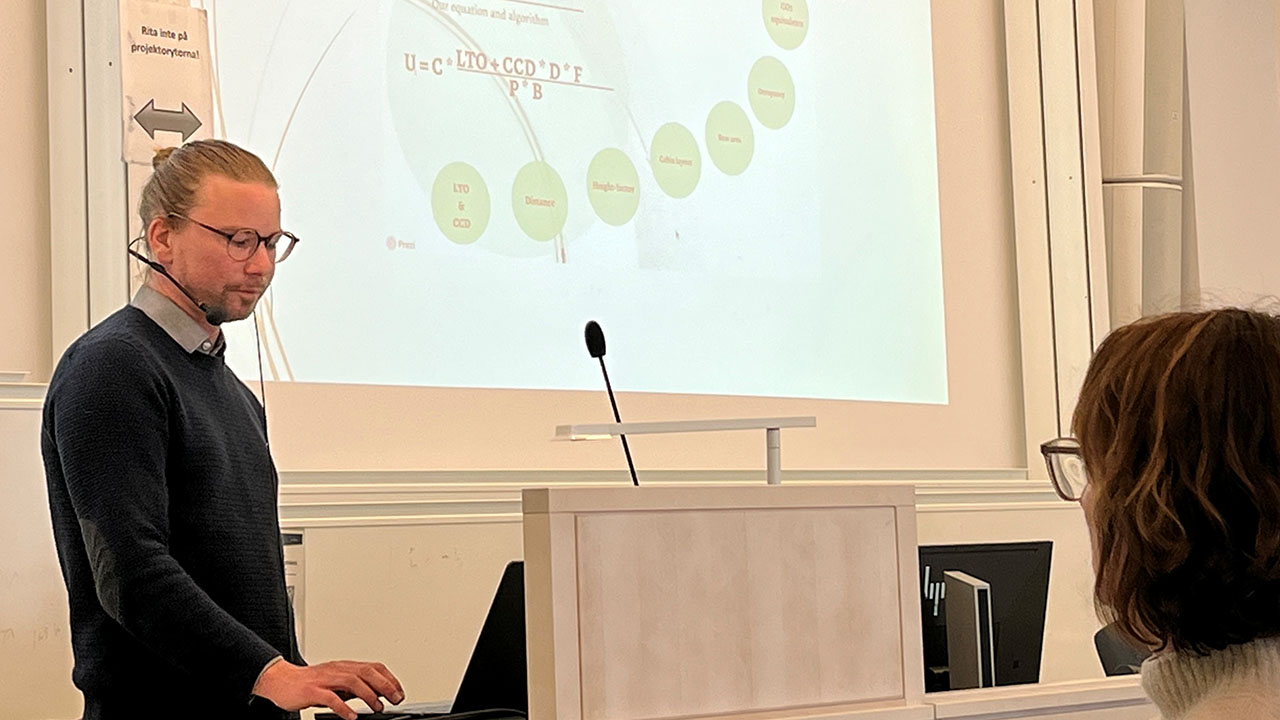 Man med håret i knut och glasögon står vid talarpulpet