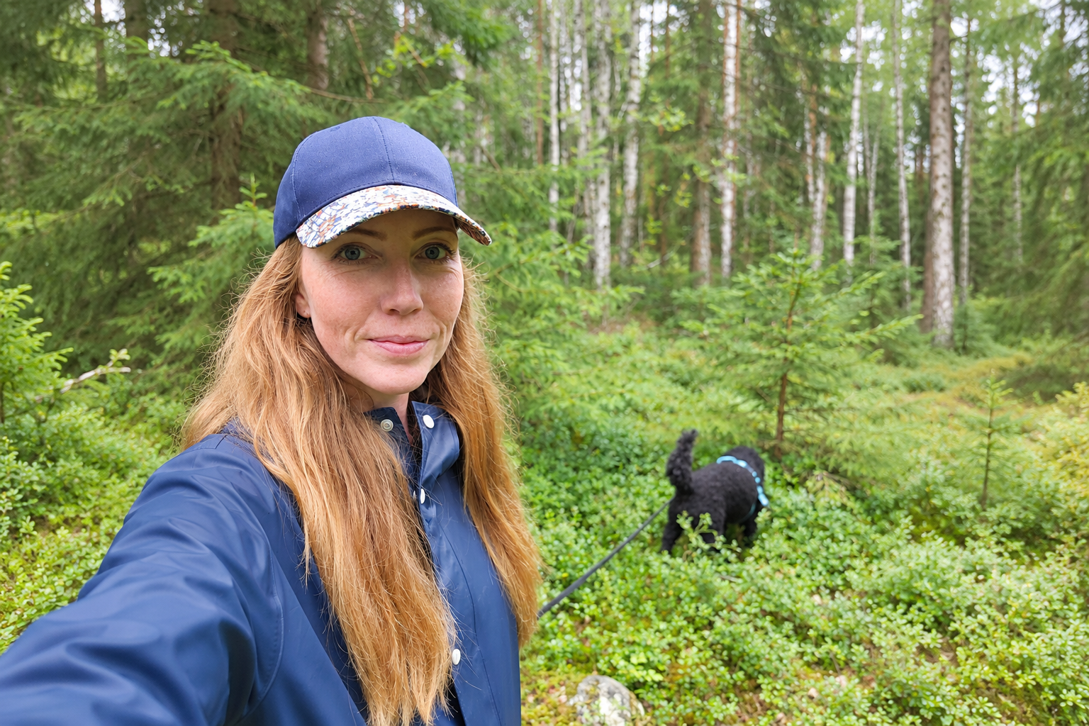 Kvinna med keps och blå jacka på promenad med en svart hund i en grönskande skog.