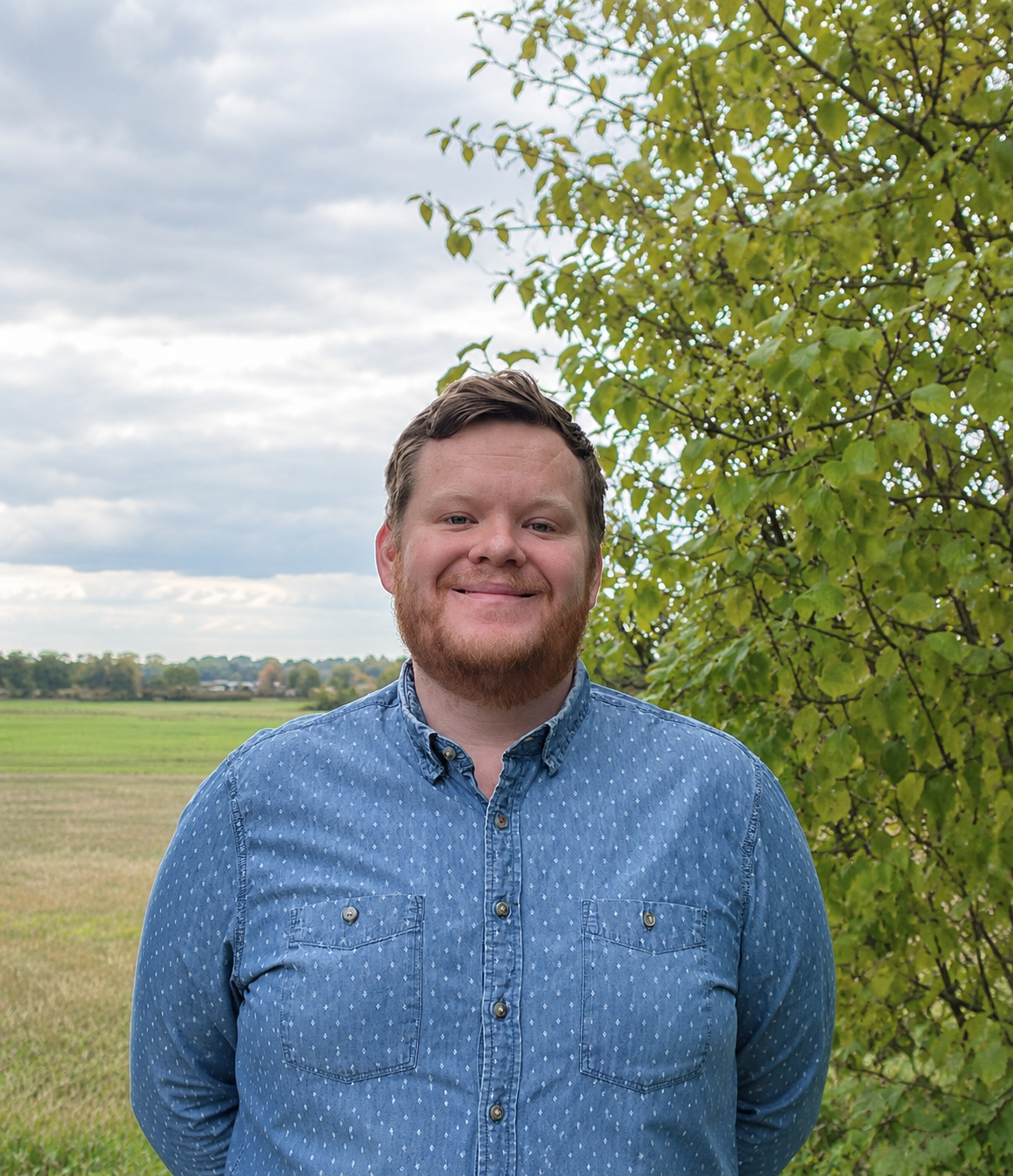 Leende man i blå skjorta framför ett grönt träd och ett öppet landskap.
