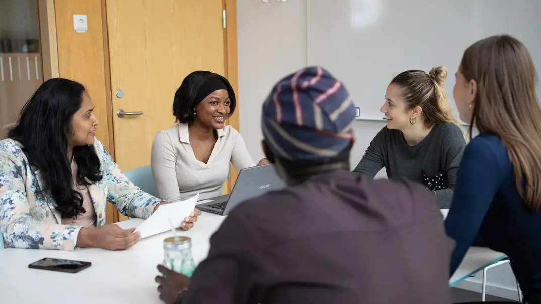 A group of five people are seated around a conference table in a meeting room, engaged in conversation and smiling. Papers, a laptop, and mobile phones are placed on the table, with a whiteboard visible in the background.