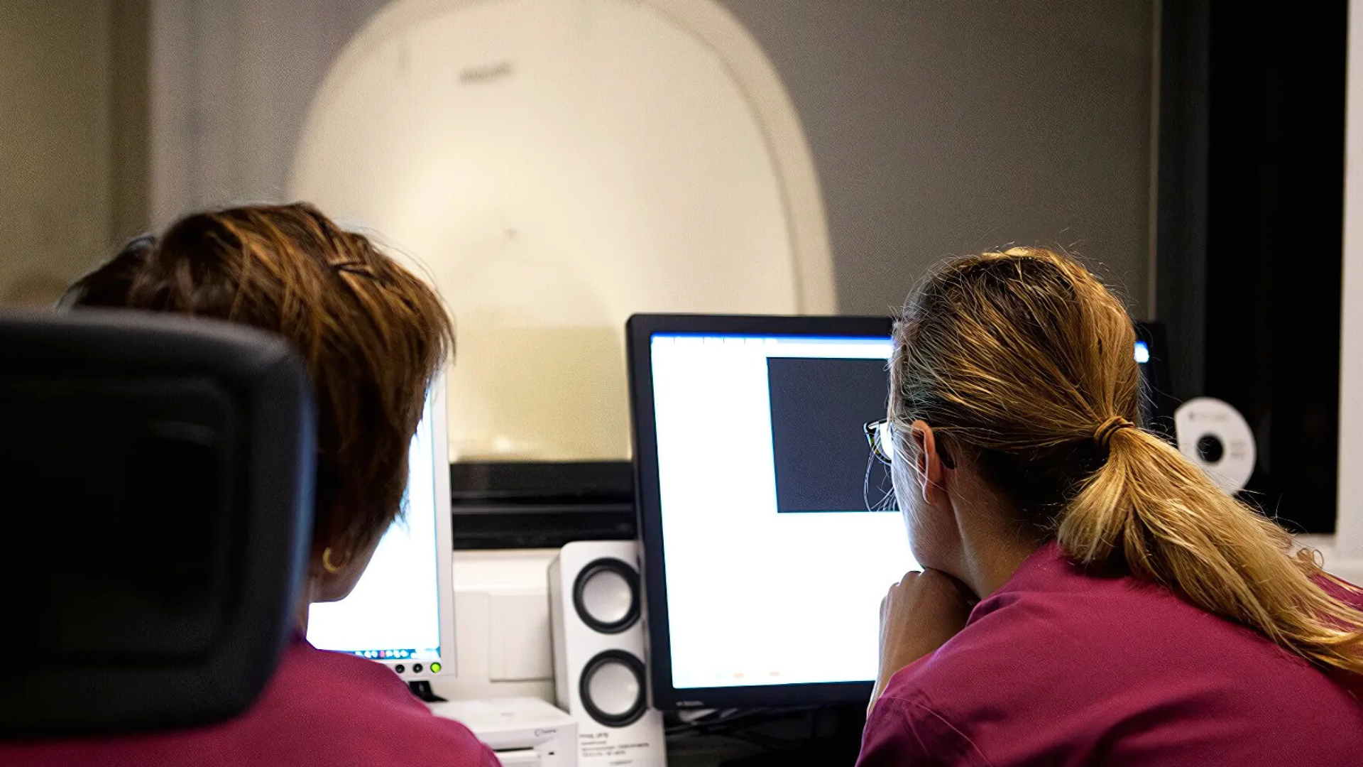 Two people sitting with their backs to the camera. One person has short brown hair and is sitting on the left side of the picture. On the right side is a woman with long blonde hair. Both people are wearing burgundy clothes. In front of the people is a computer screen that lights up white.