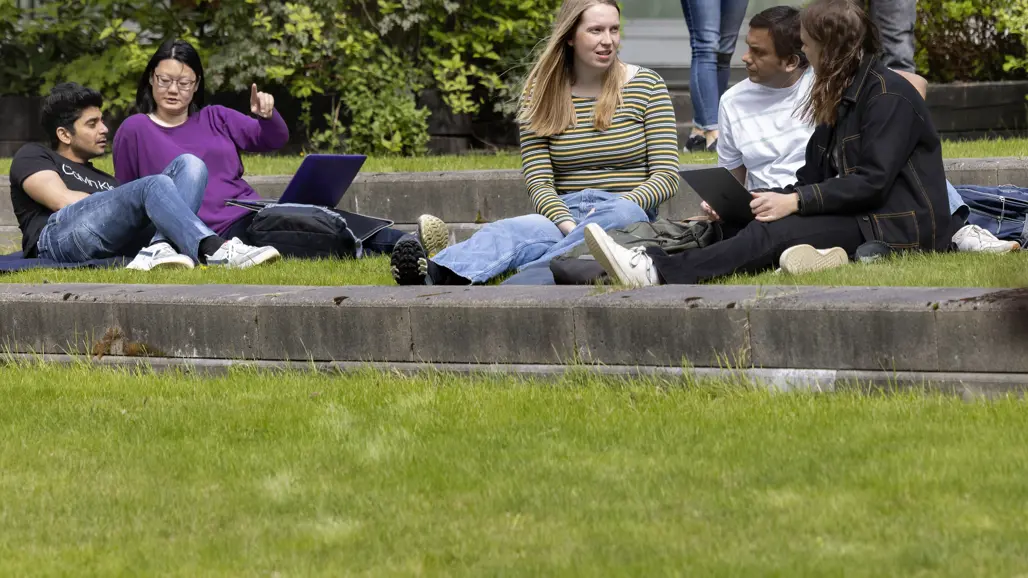 Flera studenter sitter på en gräsmatta framför en stenmur och samtalar. De har med sig laptops och ryggsäckar, och några pekar eller gestikulerar. Bakgrunden visar gröna buskar och träd under en klar dag.